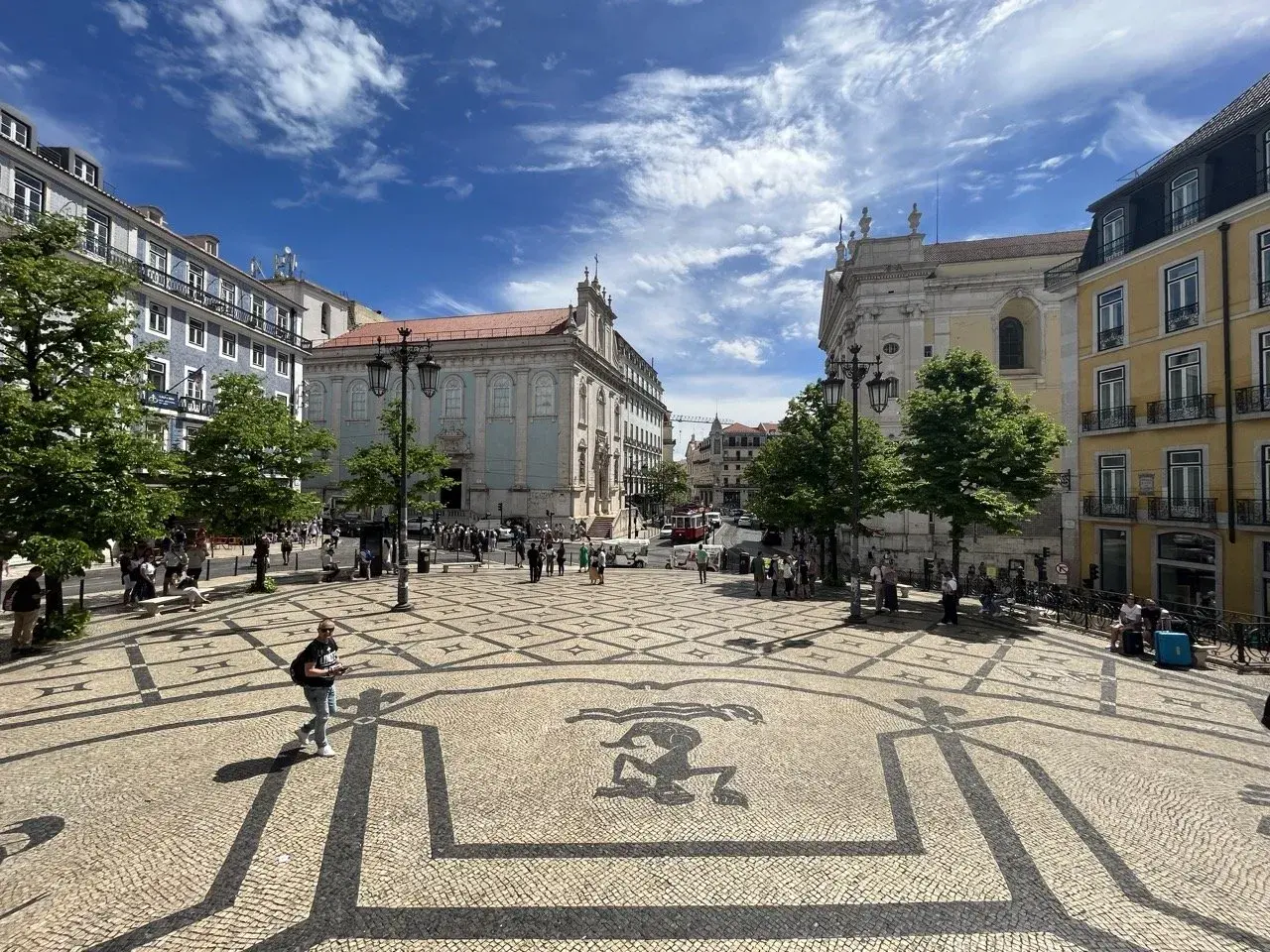A sunny day trip to Lisbon, exploring a beautiful square with intricate mosaic paving and historic buildings under a blue, cloudy sky.