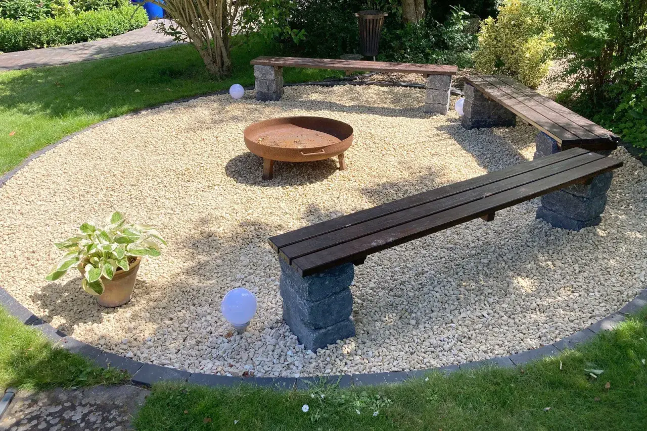 Gemütliche Kies Garten Sitzecke mit Feuerstelle und Holzbänken. Kugelleuchten sorgen für stimmungsvolles Licht.