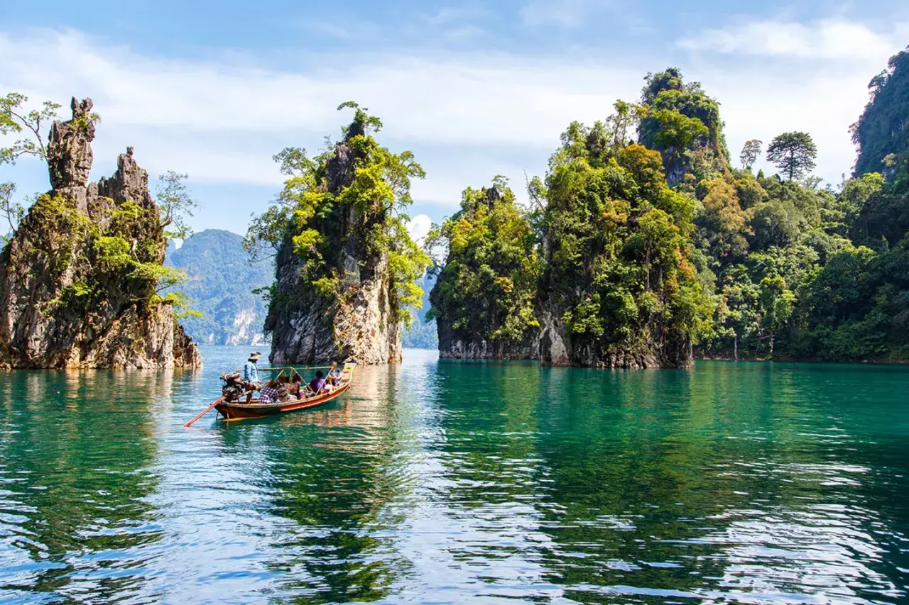 Bootfahrt durch smaragdgrünes Wasser im **Thailand Regenwald**, vorbei an steilen, bewachsenen Felsen unter blauem Himmel.
