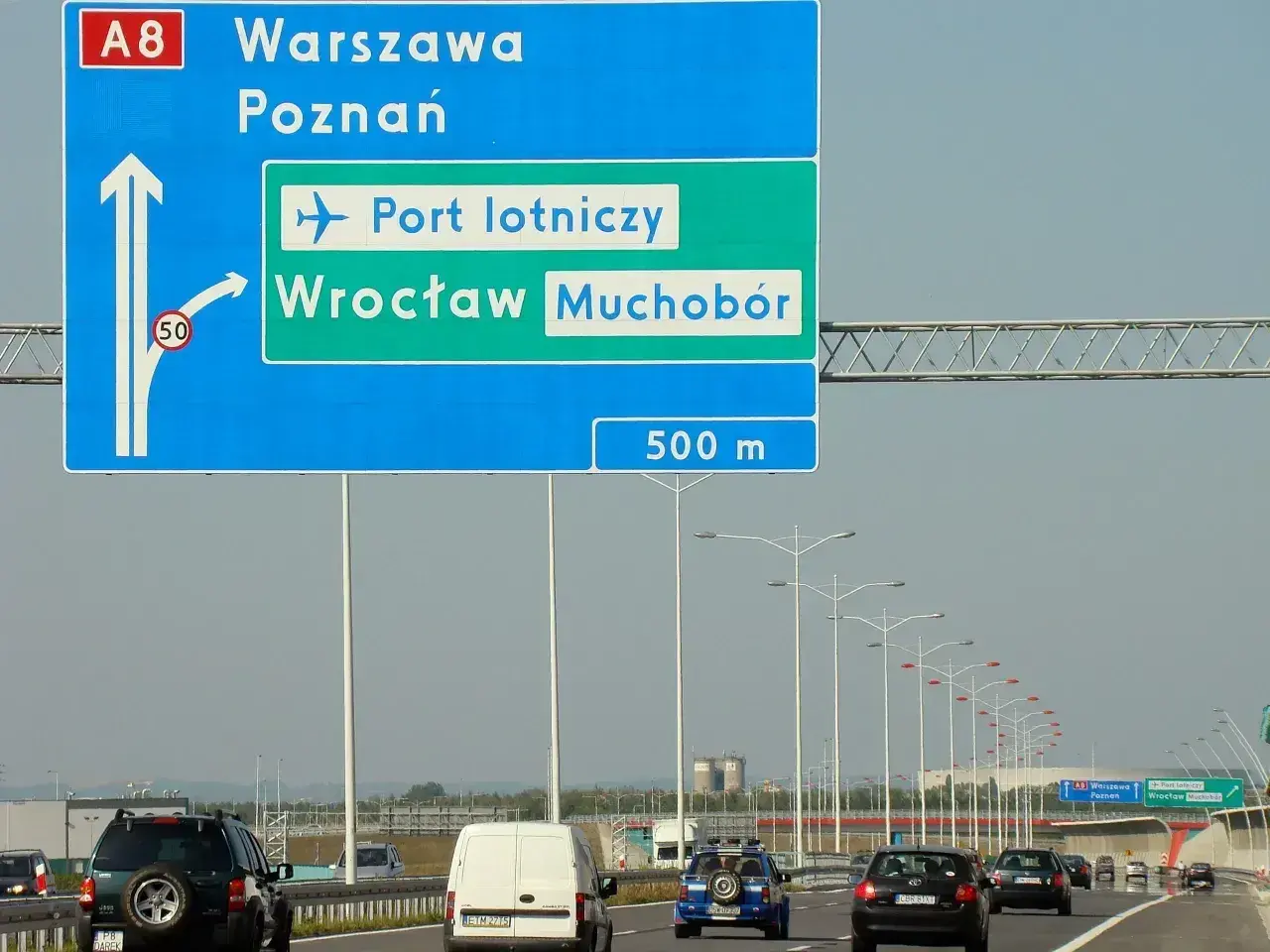 Znak drogowy pokazuje, jak dojechać na lotnisko we Wrocławiu z dworca PKP. Kierunek na lotnisko i Wrocław Muchobór jest po prawej stronie.