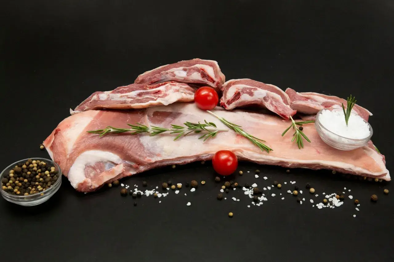 Poitrine d'agneau crue, parsemée de sel, poivre, romarin et tomates cerises, prête à être cuisinée.
