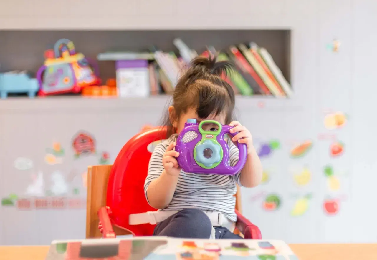 A young child in a striped shirt sits in a red high chair, holding a purple toy camera to their face.