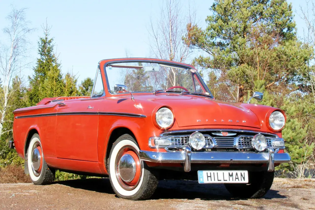 En röd cabriolet, en klassisk Hillman bil, står parkerad utomhus bland träd.