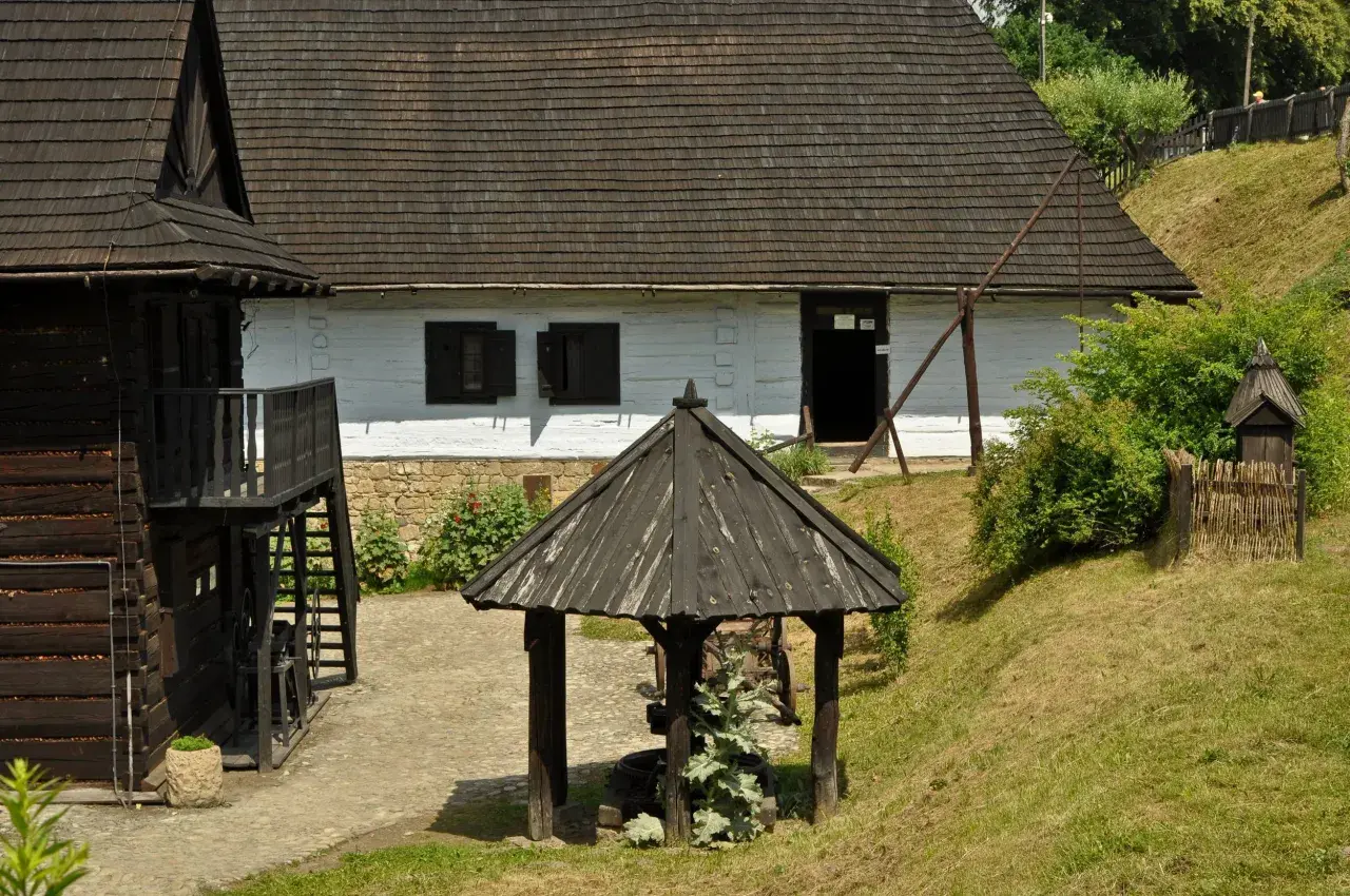 Skansen w Dobczycach: drewniane chaty, studnia i zielone wzgórza tworzą sielski krajobraz.