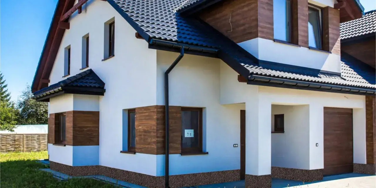Nowy dom z białym tynkiem i drewnianymi akcentami. Brak cokołu na elewacji, widoczna kamyczkowa podmurówka.