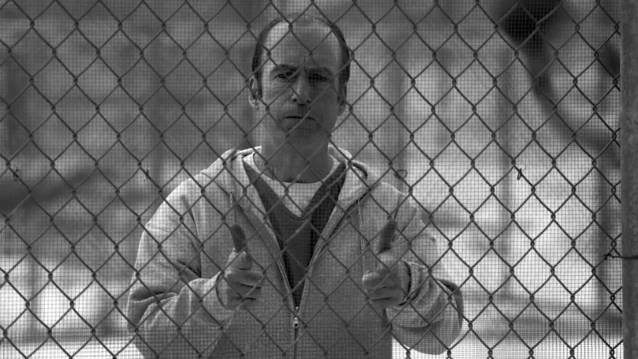 Un uomo dietro una rete metallica, con un'espressione pensierosa. Il suo gesto con i pollici in su suggerisce un "tutto bene" che contrasta con la sua situazione, evocando il **better call saul significato** di apparenza ingannevole.