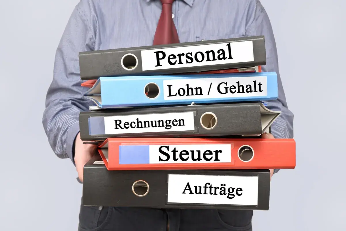 Ein Mann hält einen Stapel Ordner mit den Beschriftungen "Personal", "Lohn / Gehalt", "Rechnungen", "Steuer" und "Aufträge". Die Ordner stapeln sich und deuten auf die Komplexität der Verwaltung des zu versteuernden Einkommens hin.
