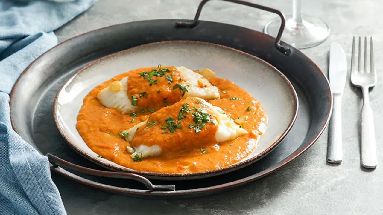Dos lomos de bacalao a la vizcaína, cubiertos de salsa roja y perejil picado, servidos en un plato sobre una bandeja metálica.