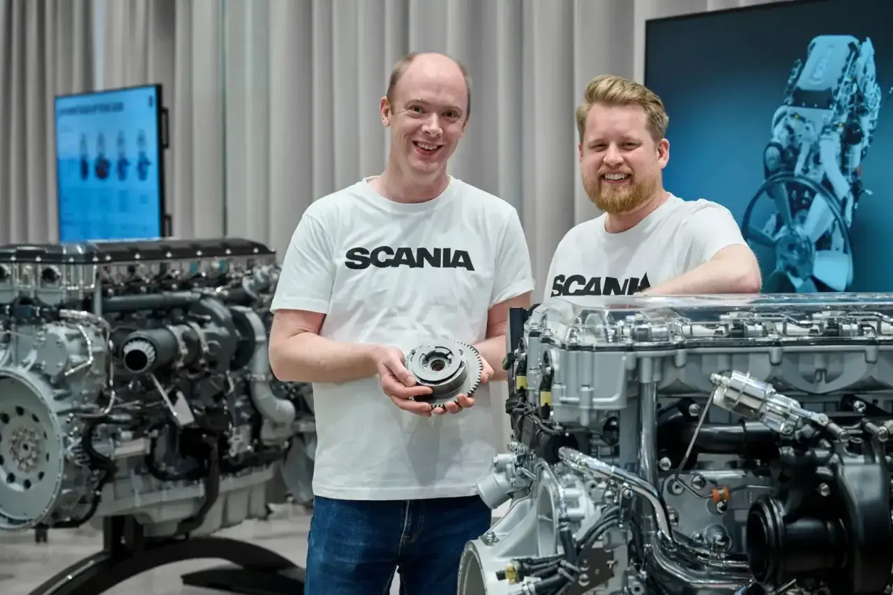 Dwóch mężczyzn w koszulkach Scania prezentuje części silnika. W tle widać potężny silnik Scania V8.