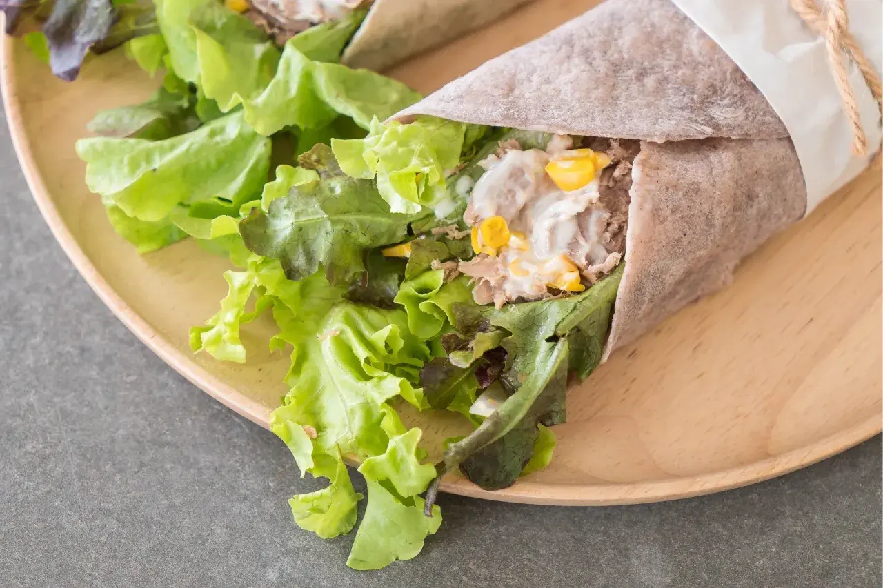 Pyszna tortilla z tuńczykiem, świeżą sałatą i kukurydzą, zawinięta w papier i przewiązana sznurkiem.
