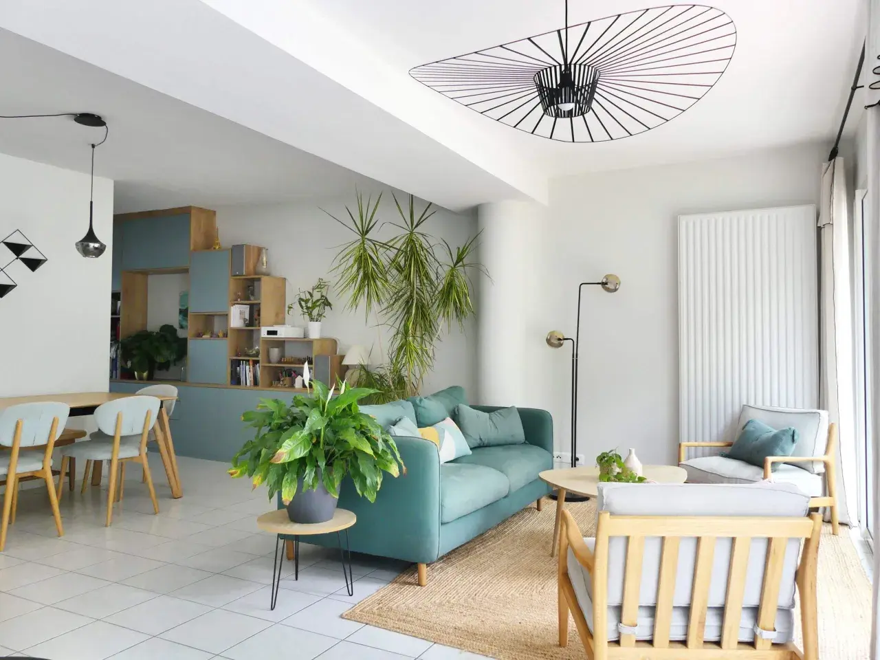 Aménagement de salon moderne avec canapé bleu sarcelle, fauteuil gris, table basse en bois et plantes vertes.