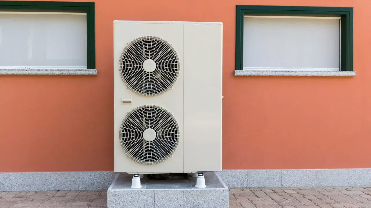 Unidad exterior de bomba de calor con dos ventiladores, instalada junto a una pared naranja con dos ventanas.