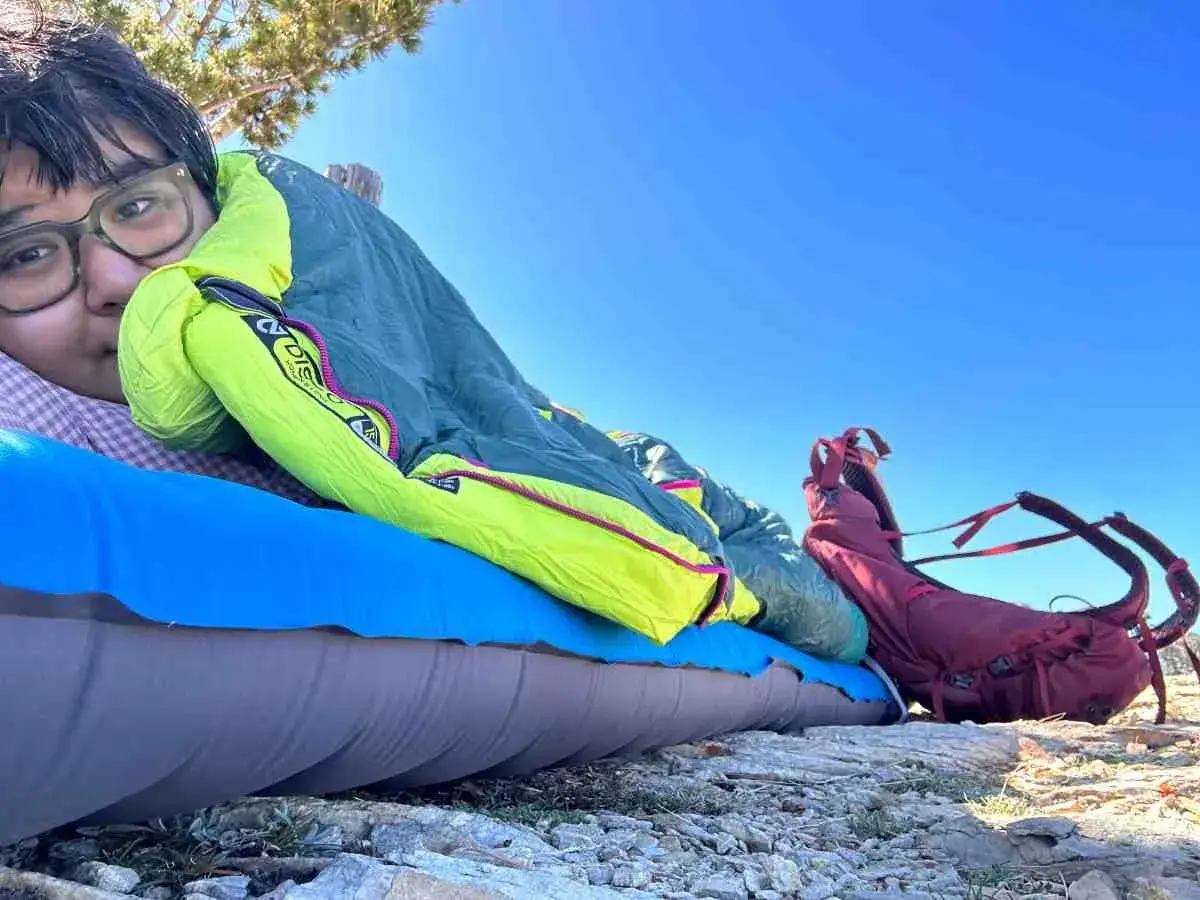 A person rests on a sleeping pad under a bright blue sky, bundled in a Nemo Disco 15 weight sleeping bag. A red backpack sits nearby.