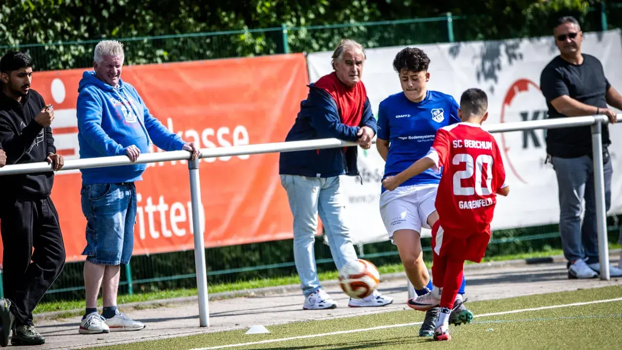 Fußball-Eltern schauen ihren Kindern beim Spiel zu. Ein Junge im roten Trikot mit der Nummer 20 spielt den Ball.