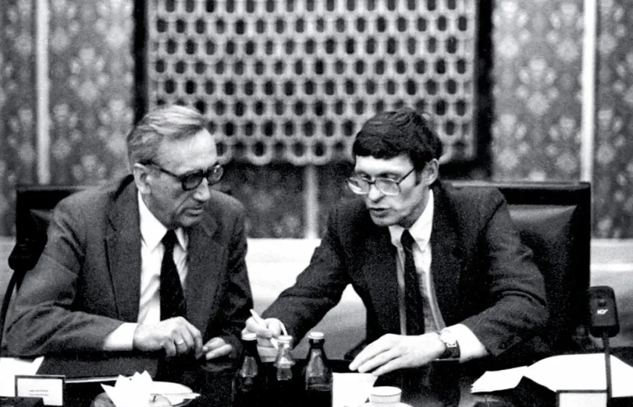 Dwóch mężczyzn w garniturach przy stole, dyskutujących o historii politycznej Polski. Antoni Dudek analizuje dokumenty.
