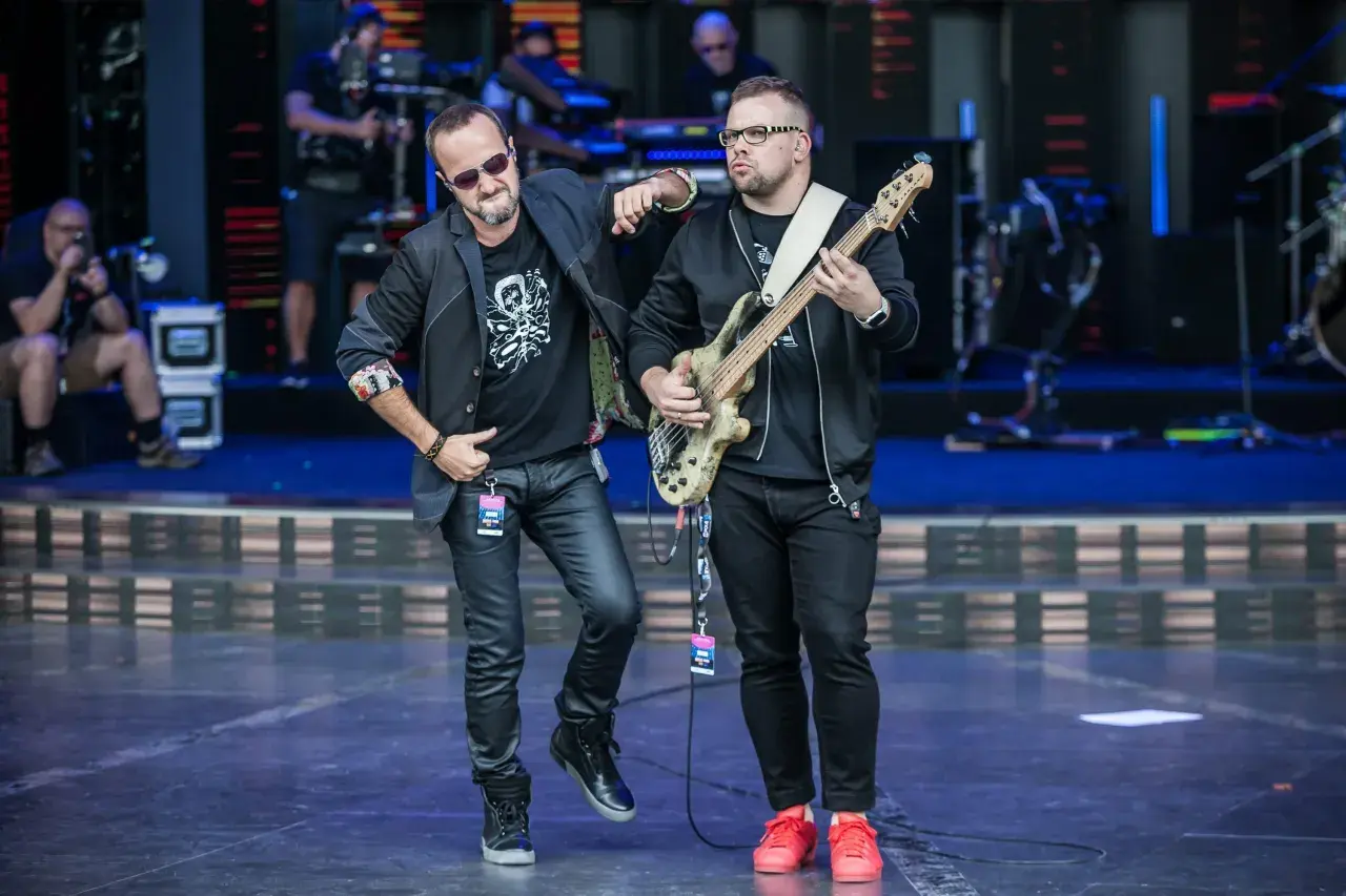 Dwóch muzyków na scenie, jeden z gitarą basową, drugi w okularach przeciwsłonecznych. Wyglądają jak nowy wokalista Kombi i jego kolega z zespołu.