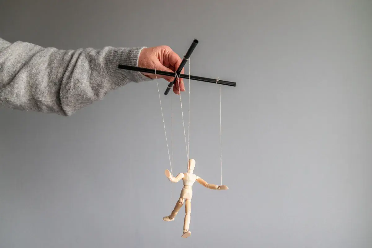 Dłoń w szarym swetrze steruje drewnianą marionetką. To obraz manipulatora, który pokazuje swoje cechy.