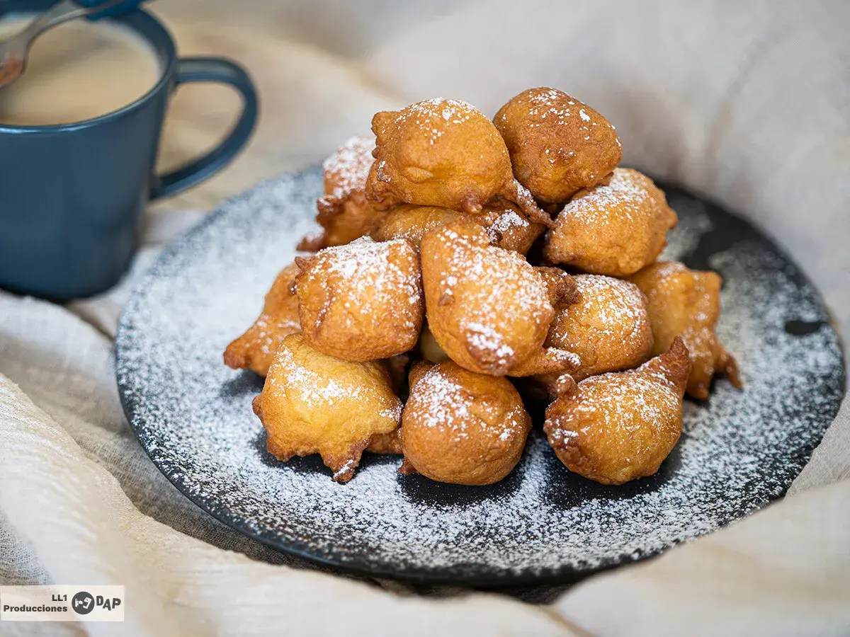Montón de buñuelos de manzana dorados y espolvoreados con azúcar glas, junto a una taza de café.