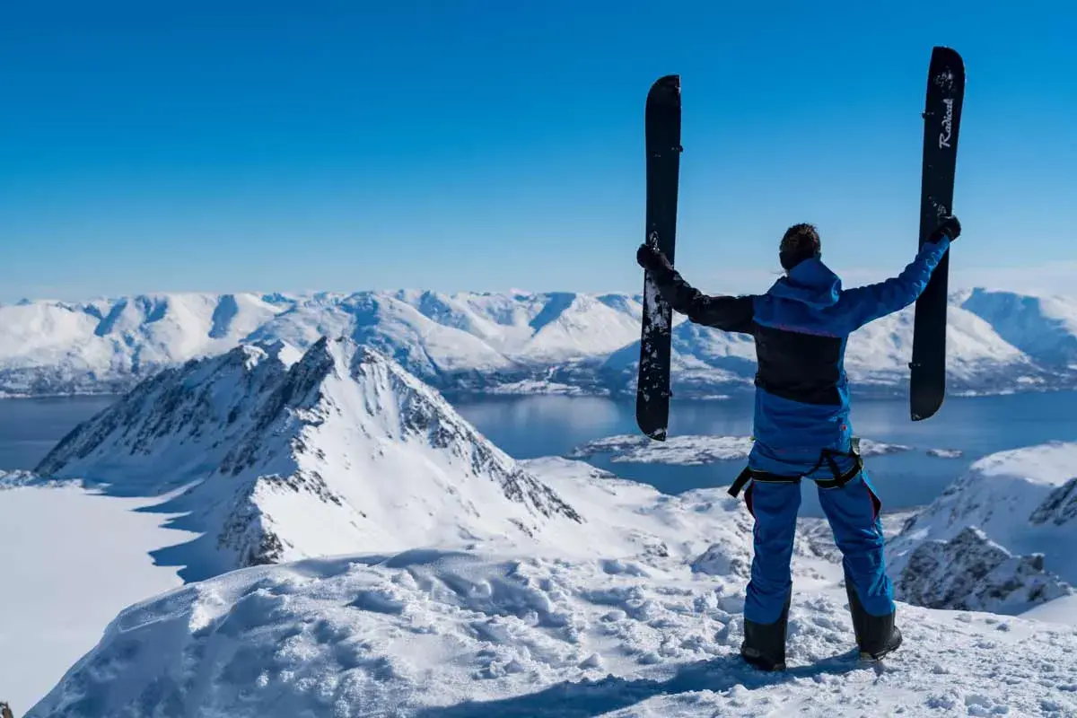 Persona en la cima de una montaña nevada, sosteniendo un splitboard que es un equipo versátil para ascender y descender.
