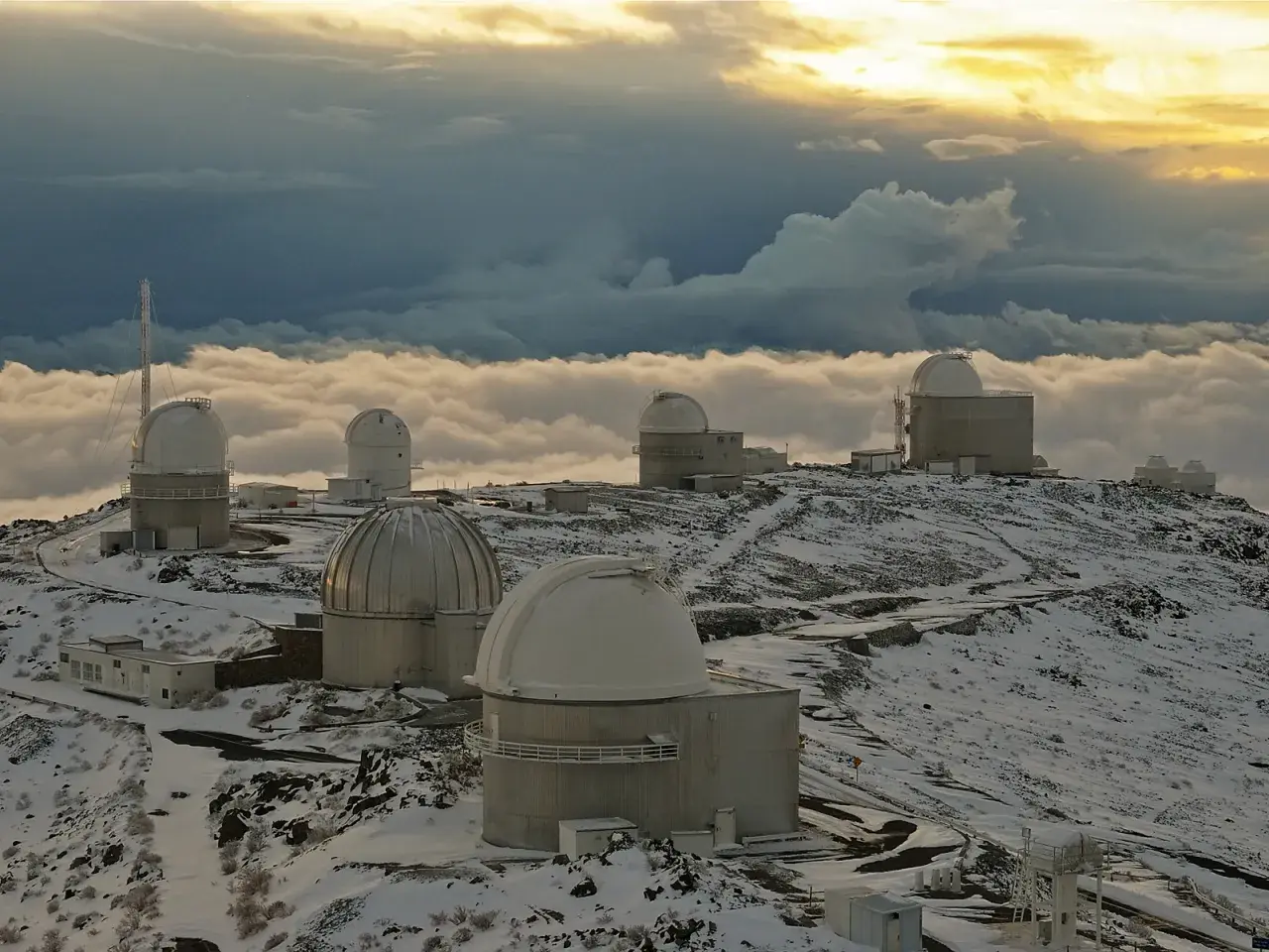 Obserwatoria astronomiczne na ośnieżonym szczycie, otoczone morzem chmur. W oddali widać złote światło zachodzącego słońca, a na pierwszym planie "la silla" - jedno z kopułowych budynków.