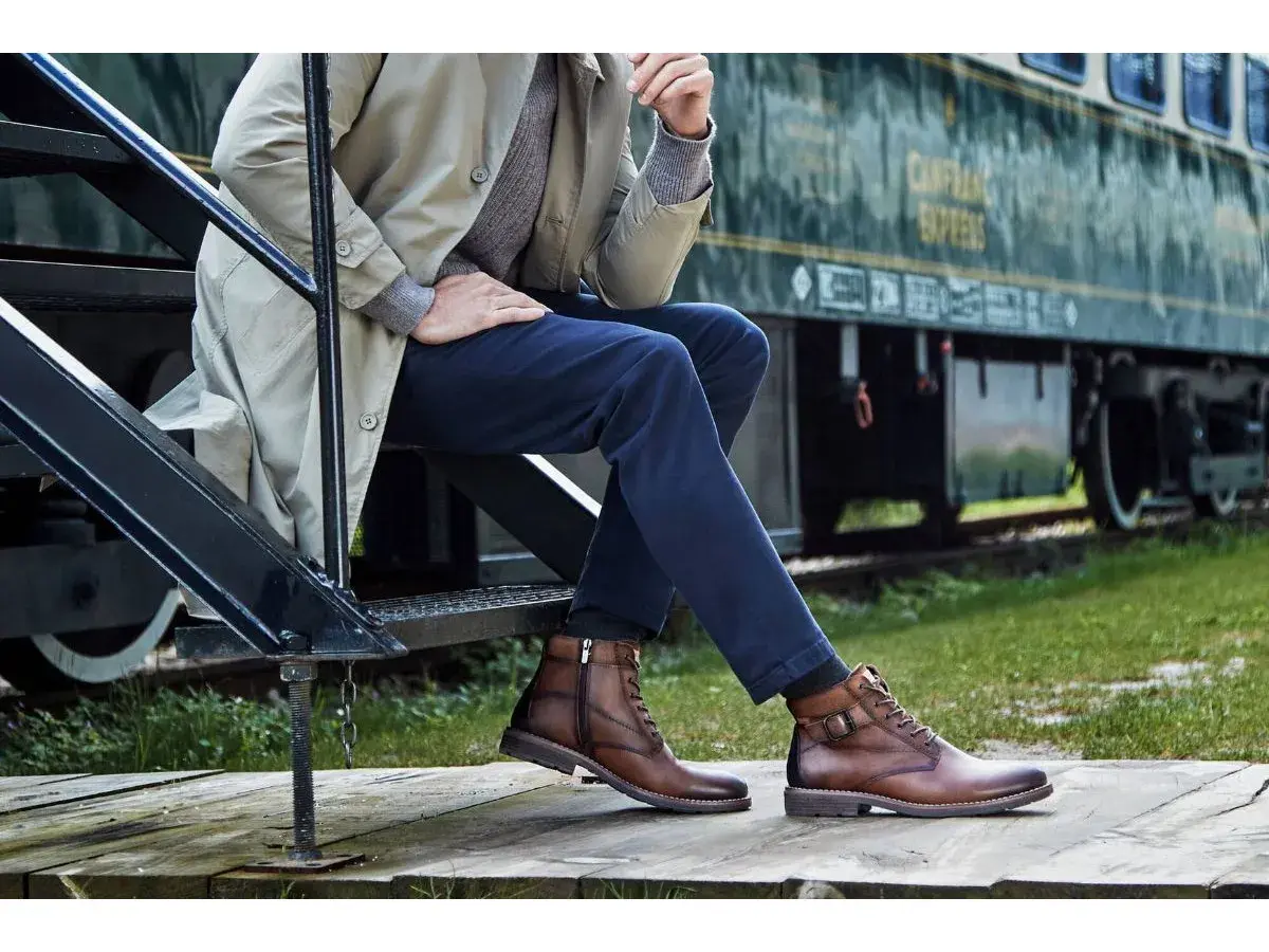Un homme élégant, assis près d'un train, porte des bottines marron. Une inspiration pour savoir quelles chaussures porter avec un jean homme.
