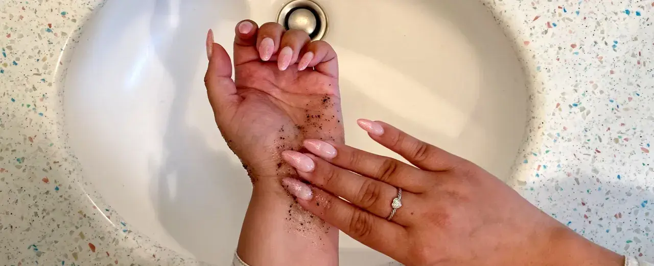 Exfoliante de café en la mano para quitar el autobronceador. Uñas largas y rosadas, anillo de corazón.
