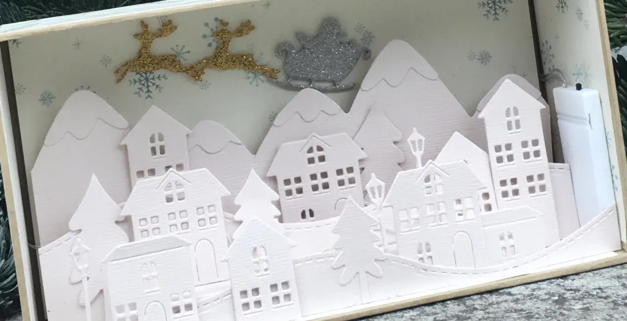 Sagome di un villaggio di Natale da ritagliare, con renne e slitta di Babbo Natale che volano sopra le casette innevate.
