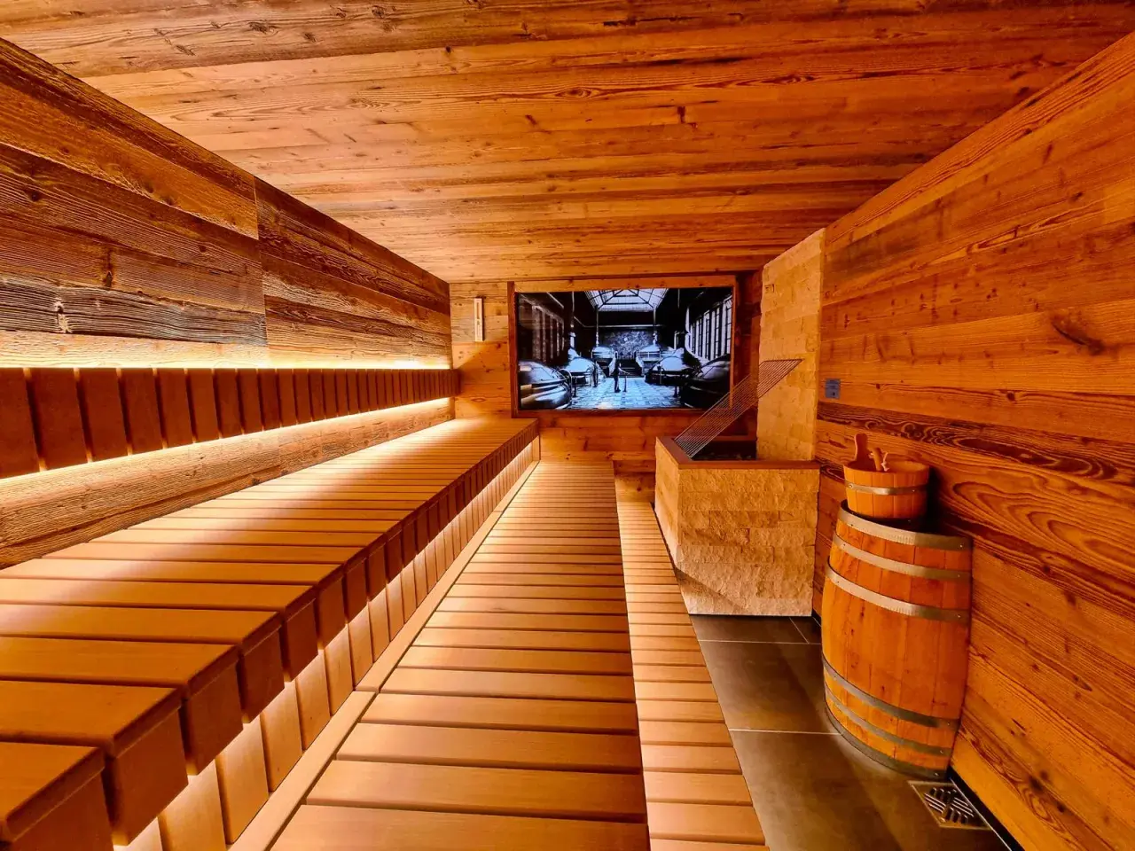 Gemütliche Holzsauna mit beleuchteten Bänken und einem Bild von einer Brauerei an der Wand. Ein Bierfass und ein Holzeimer stehen bereit für ein entspannendes Bier-Spa-Erlebnis.