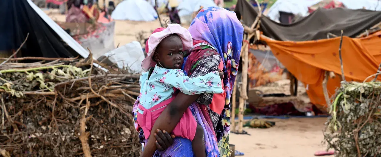 En kvinna bär ett barn i en flyktinglägerliknande miljö. Detta ger en inblick i fakta om Sudan och dess utmaningar.