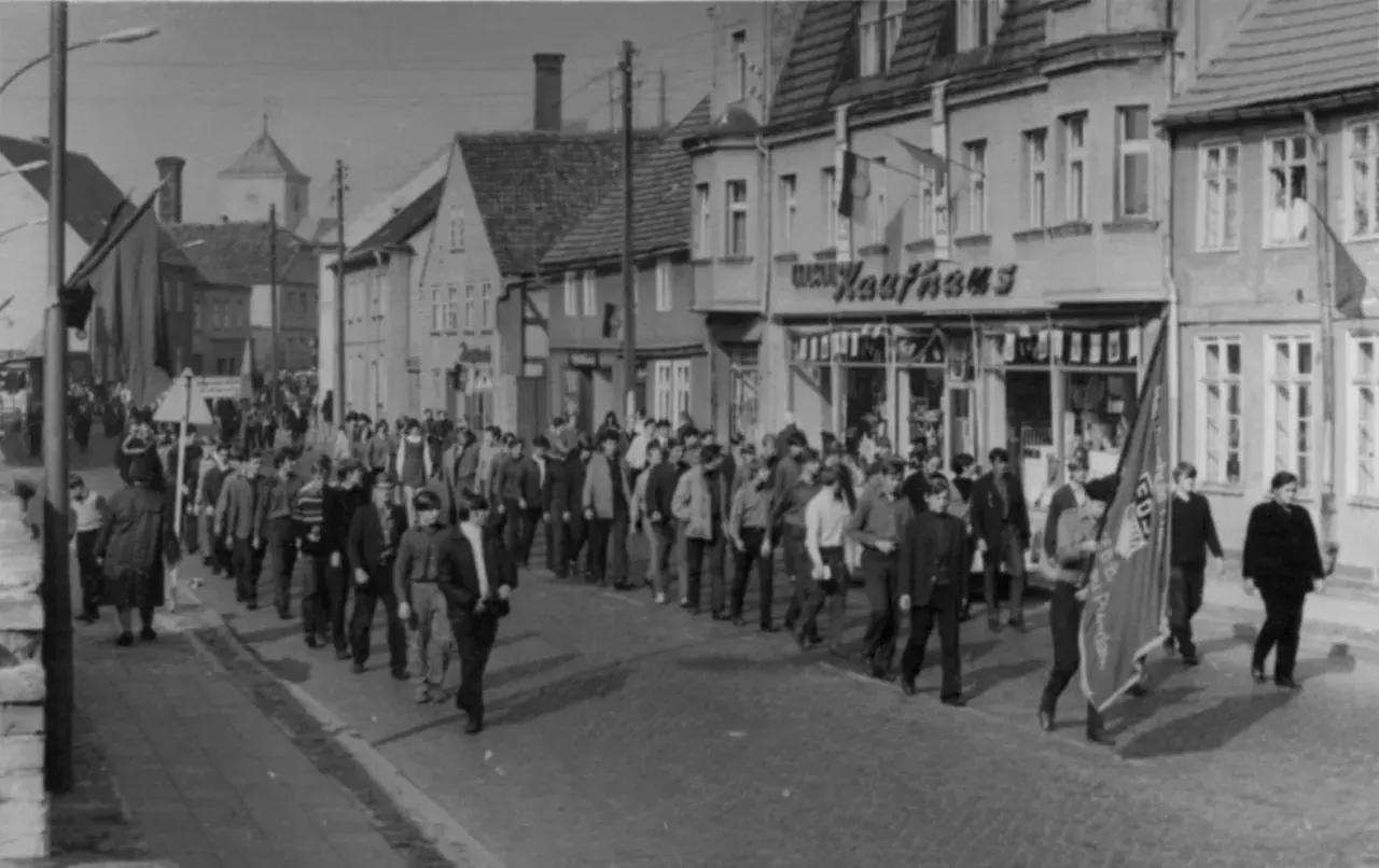 Demonstration am 7. Oktober, Tag der Republik DDR. Menschen marschieren durch eine Straße mit Geschäften, Fahnen wehen.