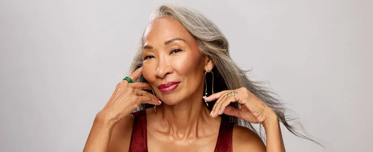 Mujer con cabello gris, con un elegante efecto de grey blending, sonríe con labios rojos y joyas doradas y verdes.