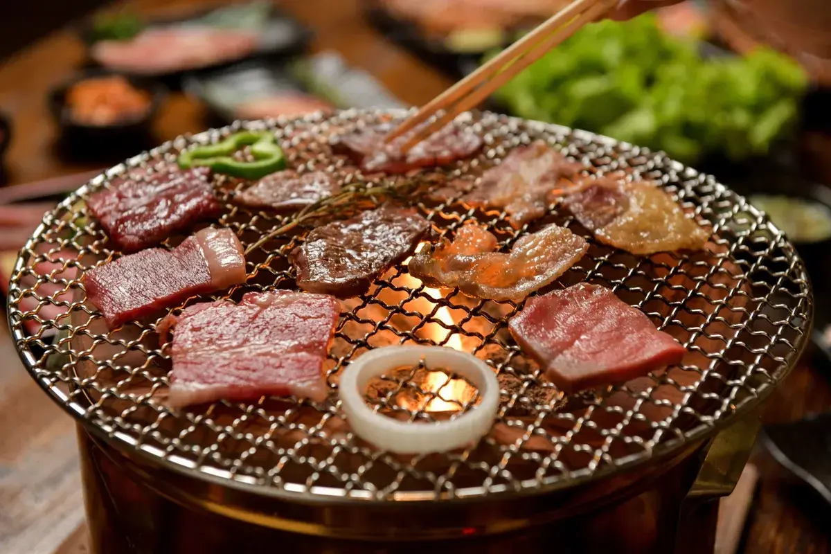 Enjoying delicious yakiniku at home. Slices of raw beef sizzle on a grill over glowing coals, with green peppers and an onion ring adding color.
