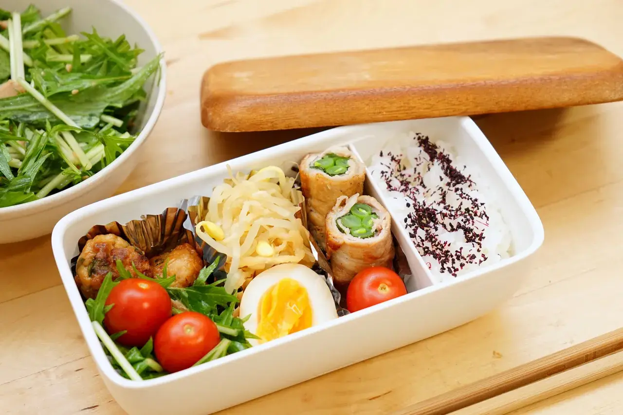 A delicious rice bento box with pork rolls, chicken meatballs, a hard-boiled egg, cherry tomatoes, and a side salad.