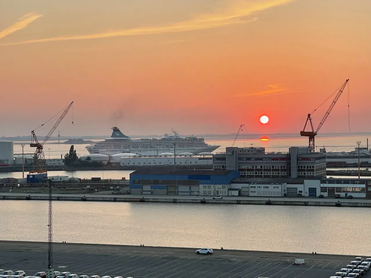 Sonnenuntergang im Hafen. Ein großes Kreuzfahrtschiff liegt vor Anker, während Kräne und Gebäude der port agency im warmen Licht erstrahlen.