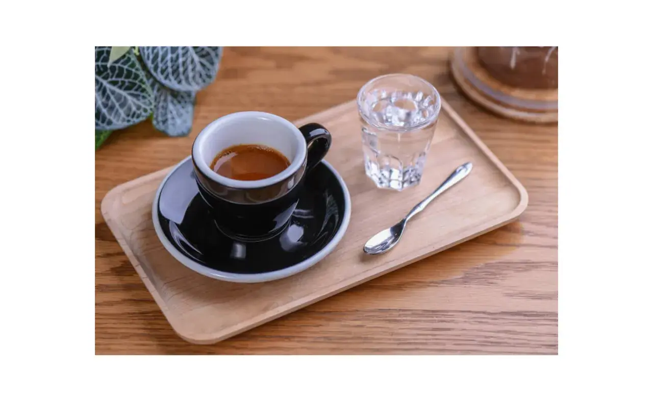 Czarna filiżanka espresso na drewnianej tacy, obok szklanka wody i łyżeczka. Idealnie pokazuje, jak podawać espresso.