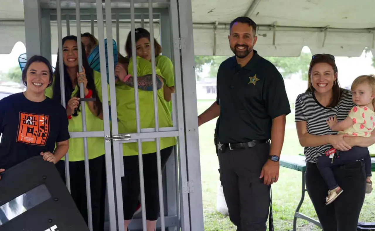 Smiles abound at a jail and bail fundraiser. A woman in a "Jail and Bail" shirt stands by a cage with women in neon shirts. A deputy and a woman holding a baby smile nearby.