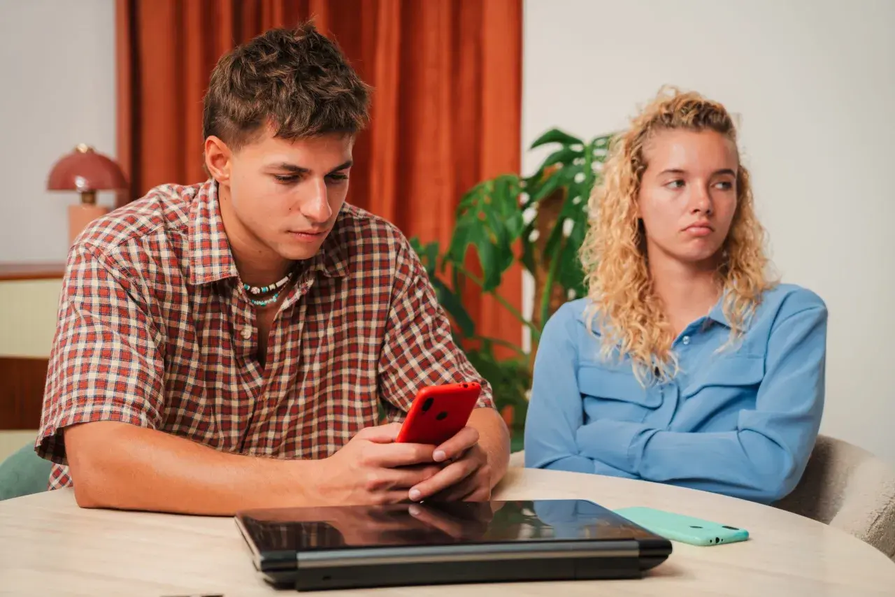 Een jongeman kijkt geconcentreerd naar zijn telefoon, terwijl zijn partner met gekruiste armen en een norse blik wegkijkt. Het voelt alsof hij haar compleet negeert, alsof ze leeggezogen wordt door zijn onoplettendheid.