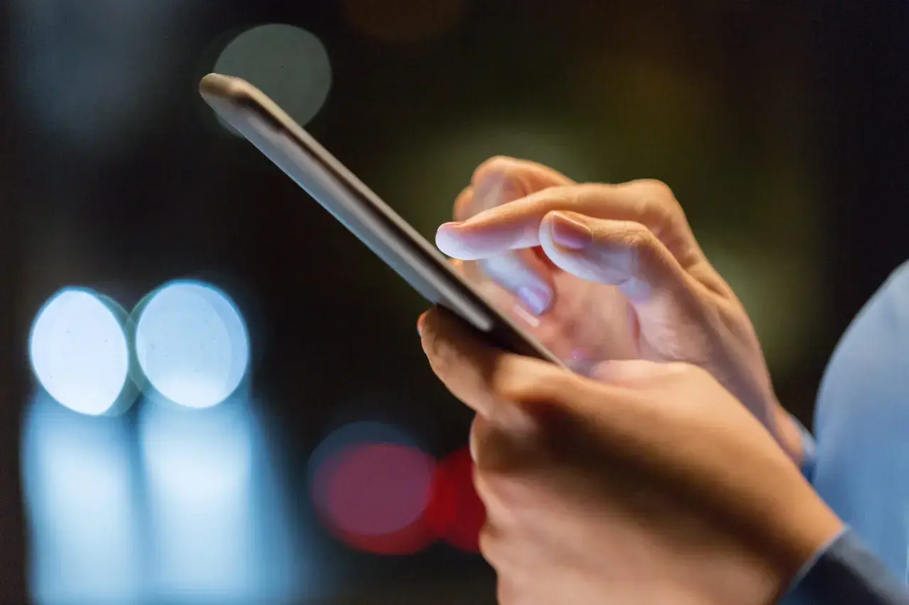 Dłoń trzyma smartfon, palec wskazujący dotyka ekranu, być może w trakcie odzyskiwania zdjęć z telefonu. Rozmyte światła w tle sugerują noc.