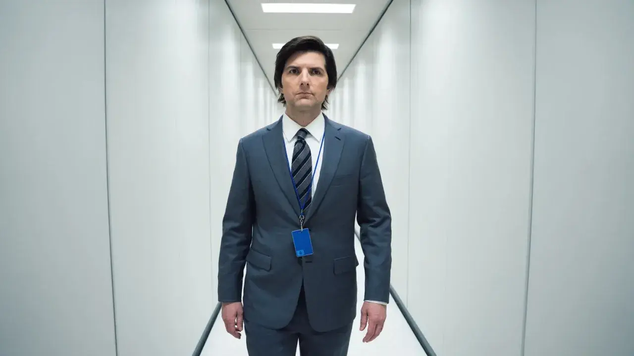 Uomo in abito grigio cammina in un corridoio bianco, con un badge blu appeso al collo. La sua espressione è seria, quasi a suggerire una scissione nella sua recensione.