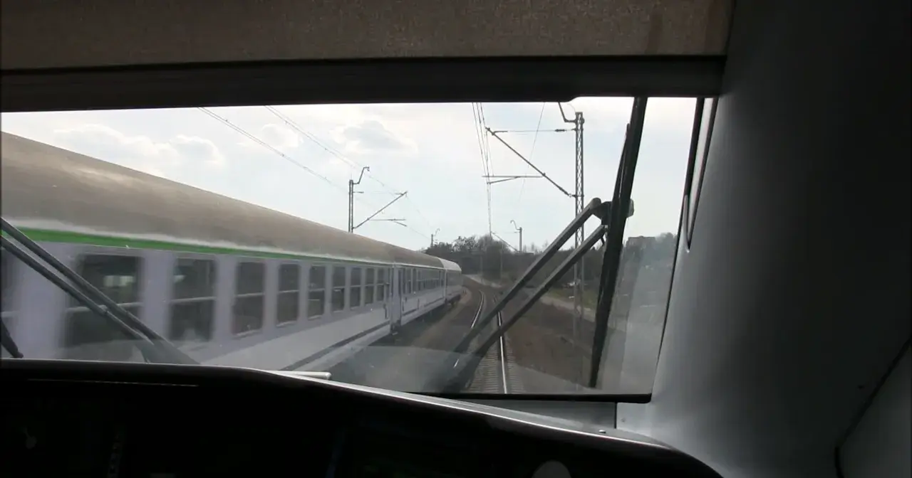 Widok z kabiny maszynisty na mijający pociąg. Widać tory, sieć trakcyjną i niebo z chmurami.