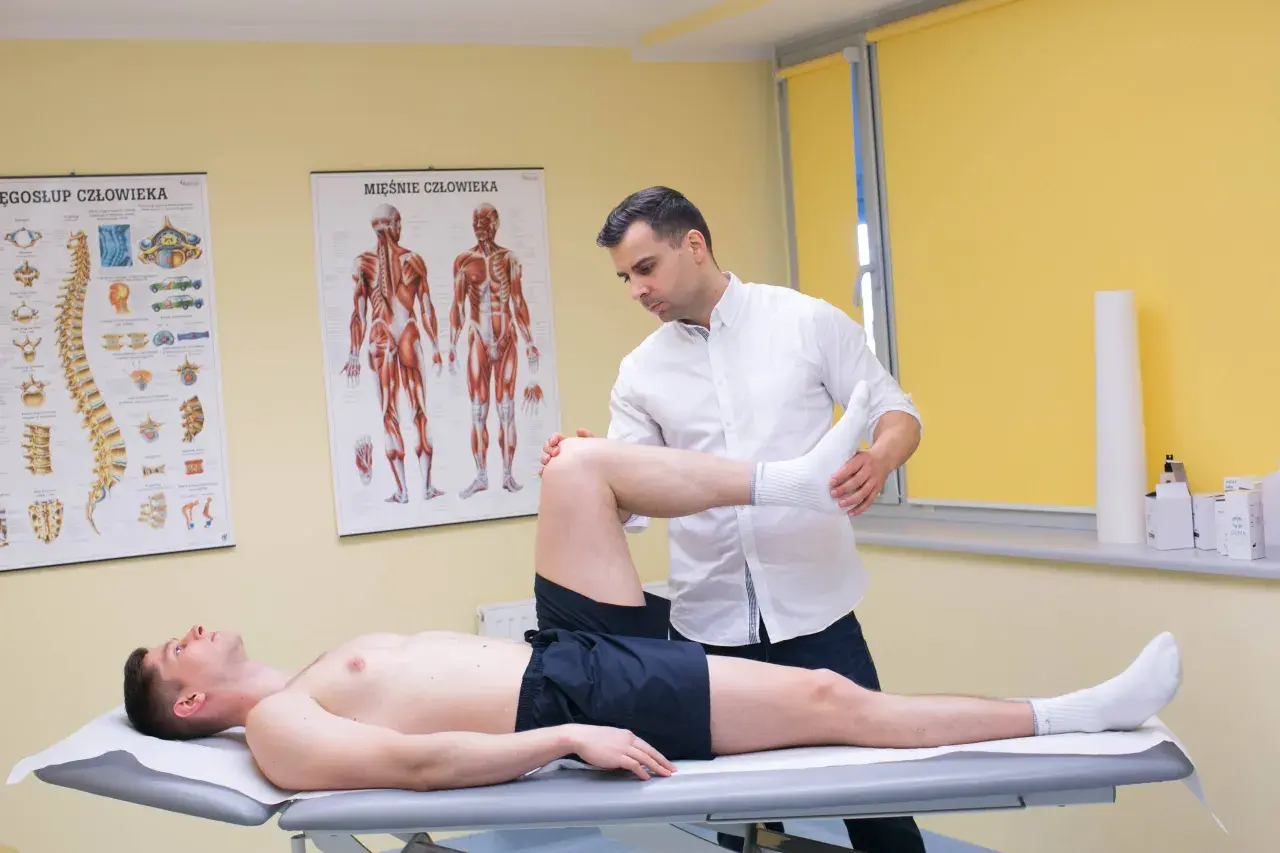 Fizjoterapeuta bada pacjenta z podejrzeniem naderwanego ścięgna. Na ścianie wiszą anatomiczne plakaty.