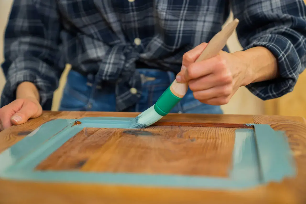 Person streicht mit Pinsel und hellblauer Farbe über Holz. Ein DIY-Projekt, um alten Möbeln neuen Glanz zu verleihen. Lack auf Lack streichen.