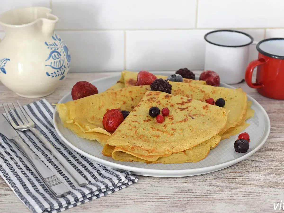 Deliciosos crepes de avena doblados y cubiertos con bayas congeladas y frescas. Servidos en un plato blanco con cubiertos y una jarra de cerámica al fondo.