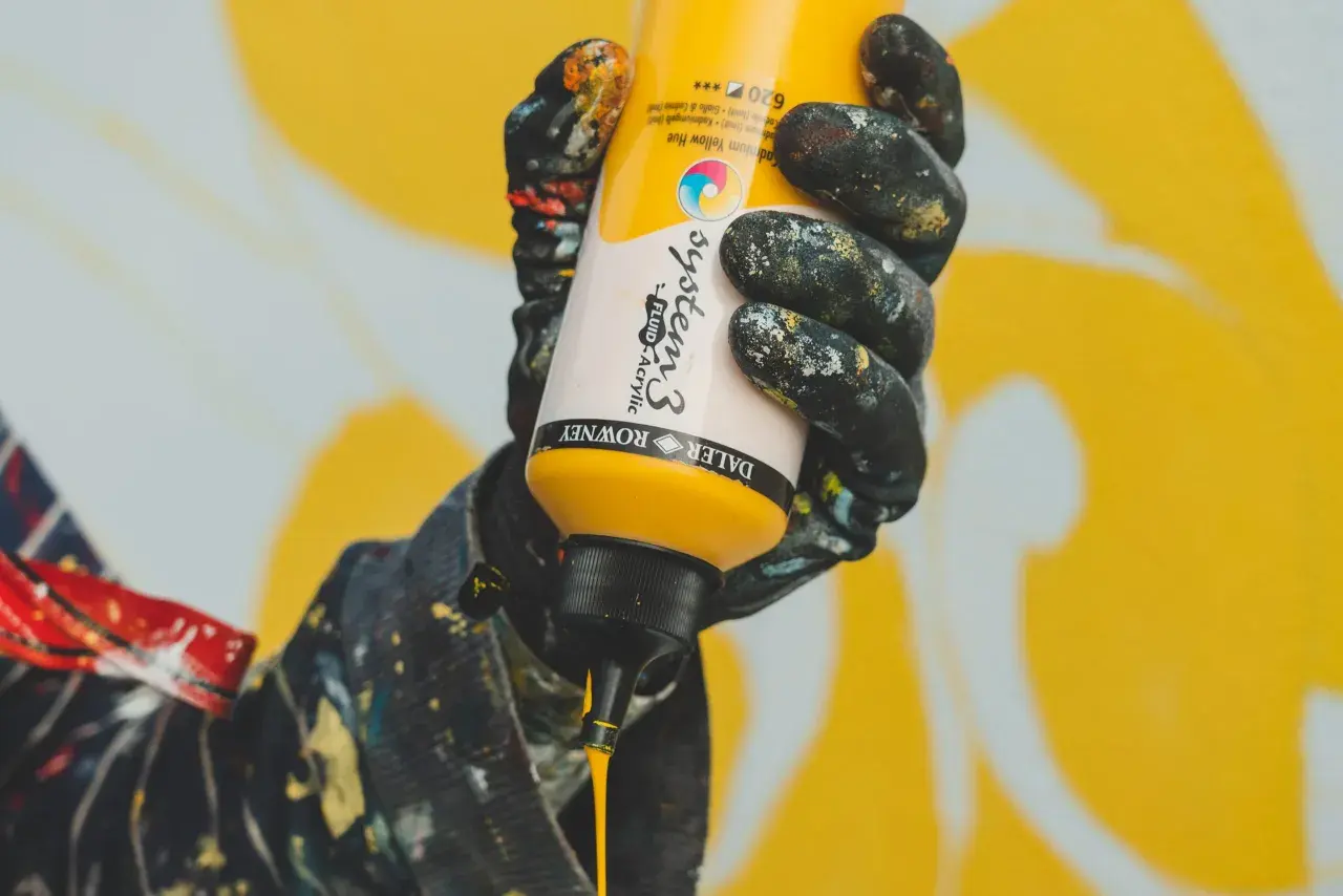 Mano con guanti sporchi di vernice che stringe un flacone di giallo brillante, pronto a creare un'opera d'arte. Il colore viola è assente, ma l'energia creativa è palpabile.