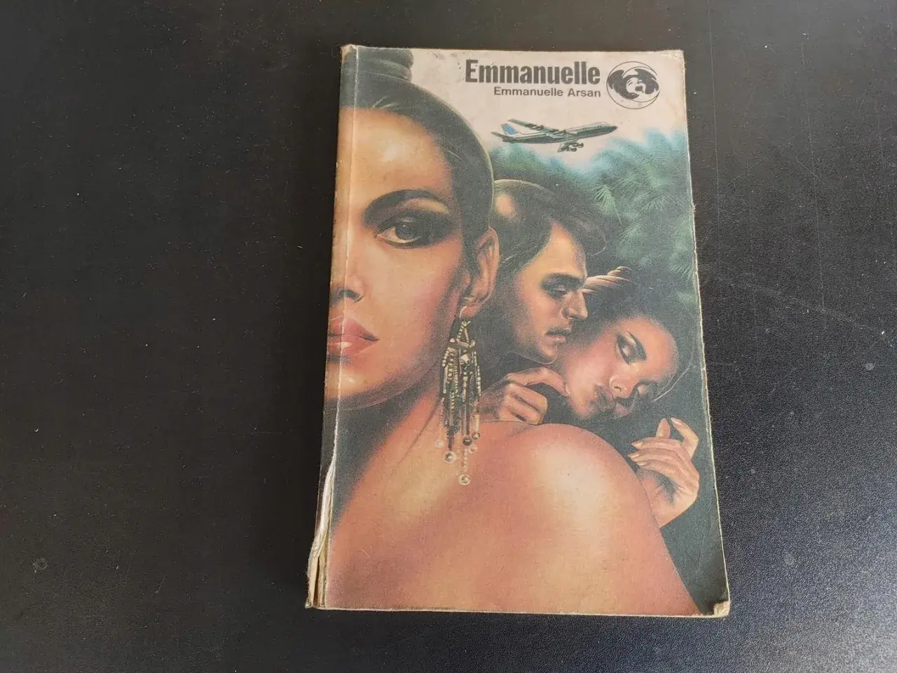 Okładka książki "Emmanuelle" z wizerunkiem kobiety i pary, w tle samolot i dżungla.