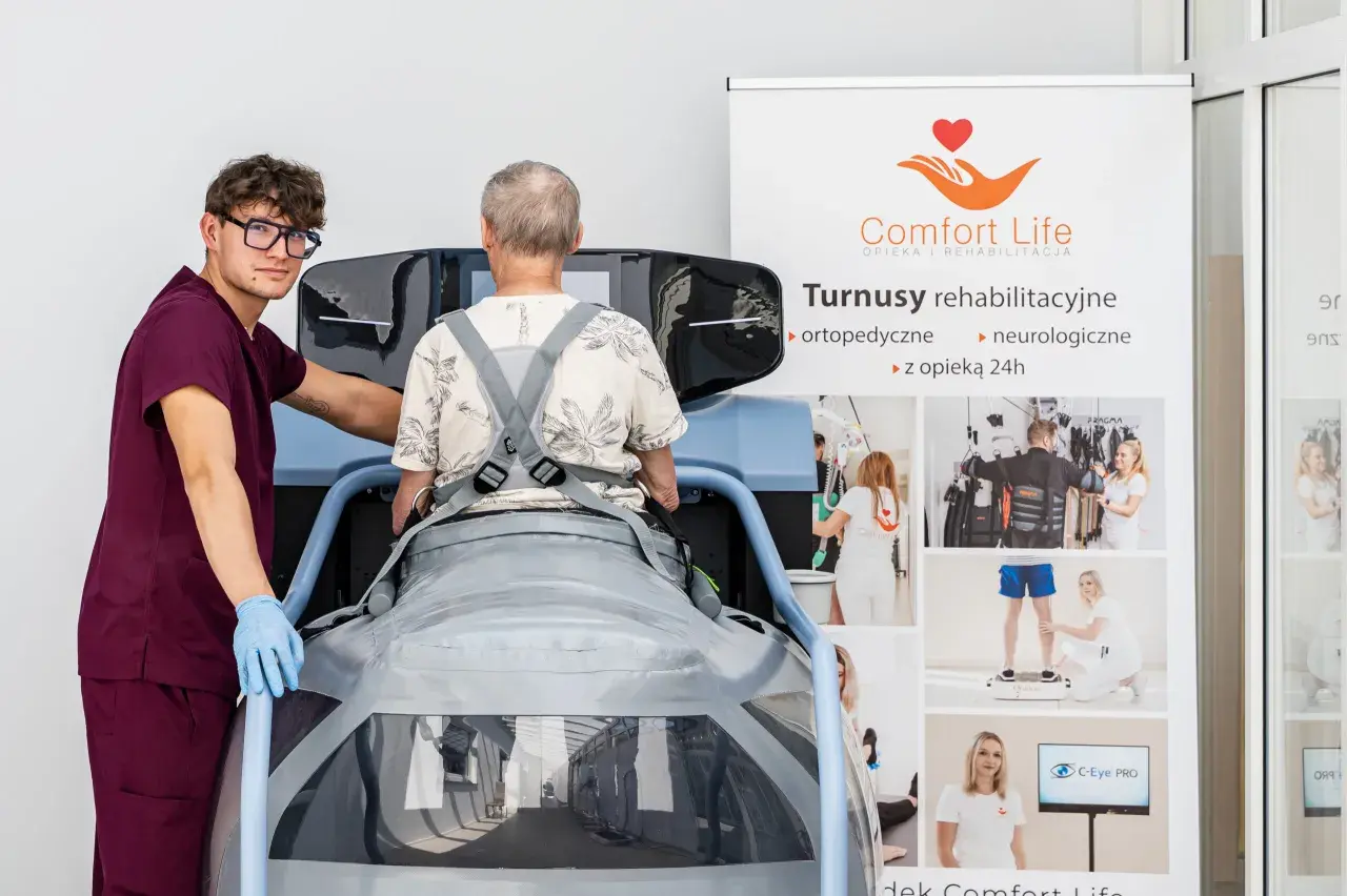 Rehabilitacja po operacji kręgosłupa: pacjent w specjalistycznym urządzeniu, asystuje mu terapeuta. Na banerze informacje o turnusach.
