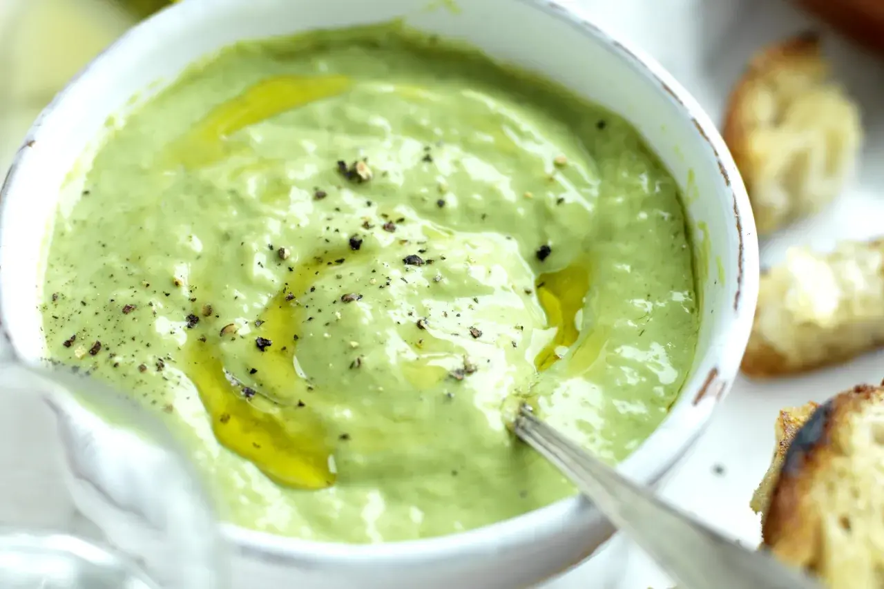 A bowl of creamy green basil pesto with olive oil and cracked pepper, served with toasted bread.
