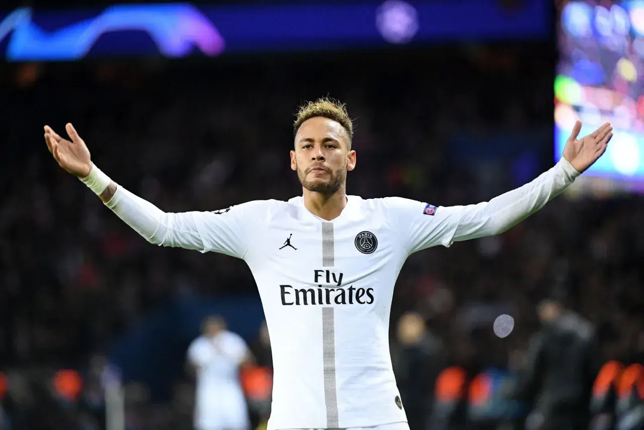 Neymar w białej koszulce PSG, z charakterystycznymi fryzurami, unosi ręce na tle tłumu.
