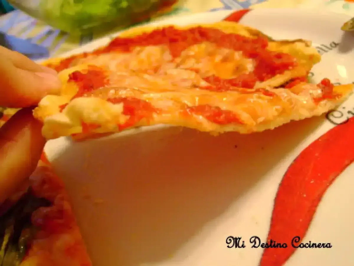 Una mano sostiene una rebanada de pizza con queso derretido y salsa de tomate, sobre un plato decorado con pimientos rojos. La masa, hecha con semola de trigo, luce dorada y crujiente.