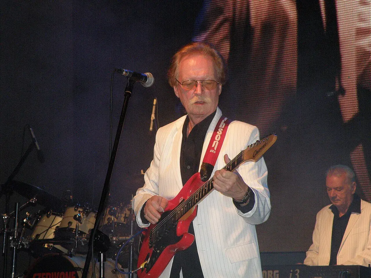 Muzyk w białej marynarce gra na czerwonej gitarze. Na scenie widać też perkusję i klawisze.